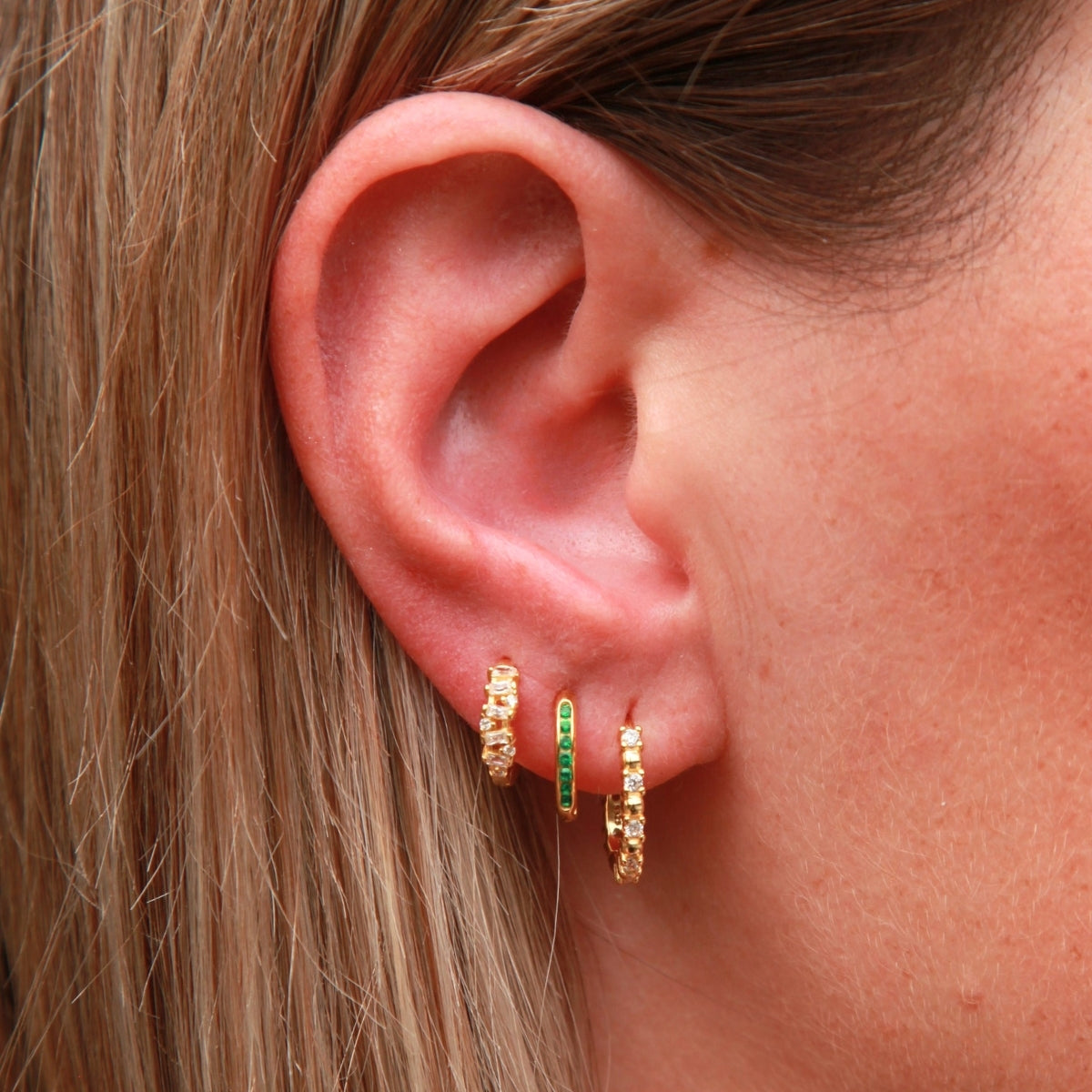 Close-up of an ear wearing gold hoop earrings with green gemstones.