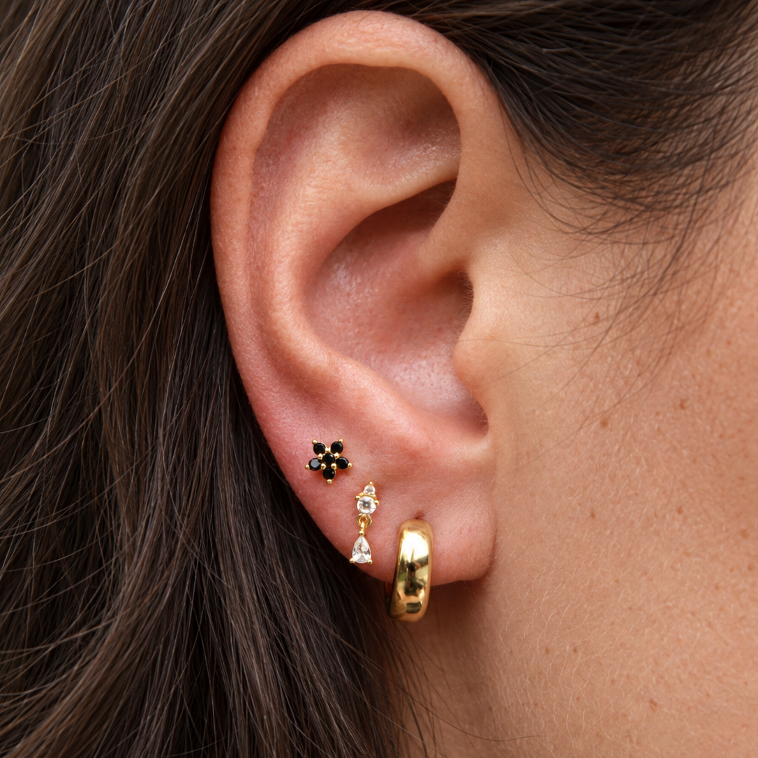 Close-up of a person's ear showcasing multiple gold and black stud earrings, including a small black flower-shaped stud, a tiny gold and white bead drop, and a textured gold huggie hoop.