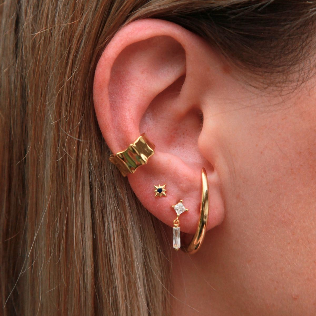 Close-up of an ear wearing gold earrings and structural wide cuff with a blurred background