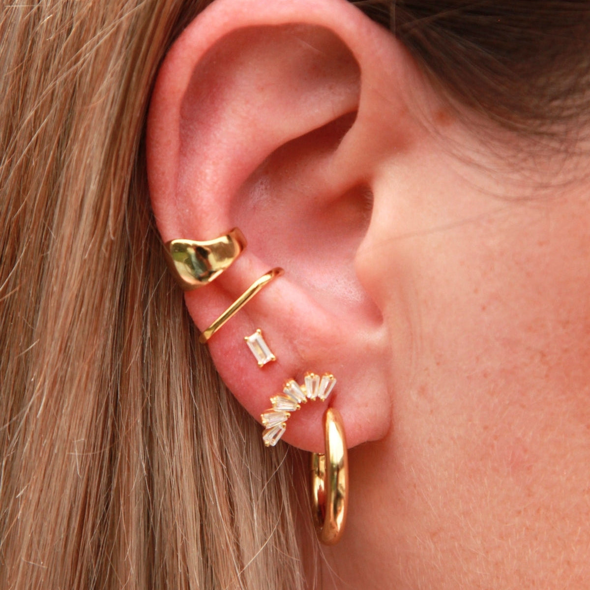 Close-up of an ear with gold earrings stacked and blonde hair. One of the earrings crown-like shape with baguette-cut stones