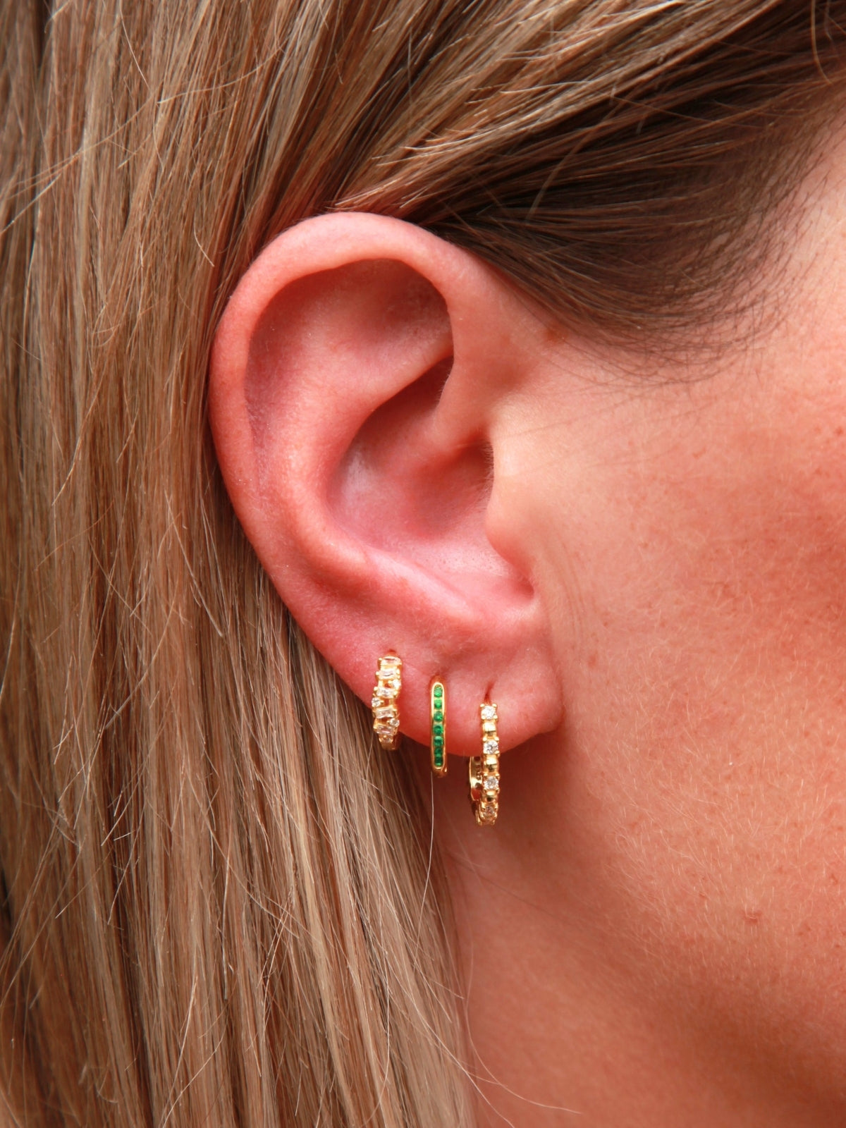 Close-up of an ear wearing gold hoop earrings with green gemstones.