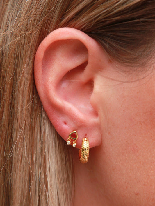 Close-up of an ear wearing gold hoop earrings with a red gemstone.