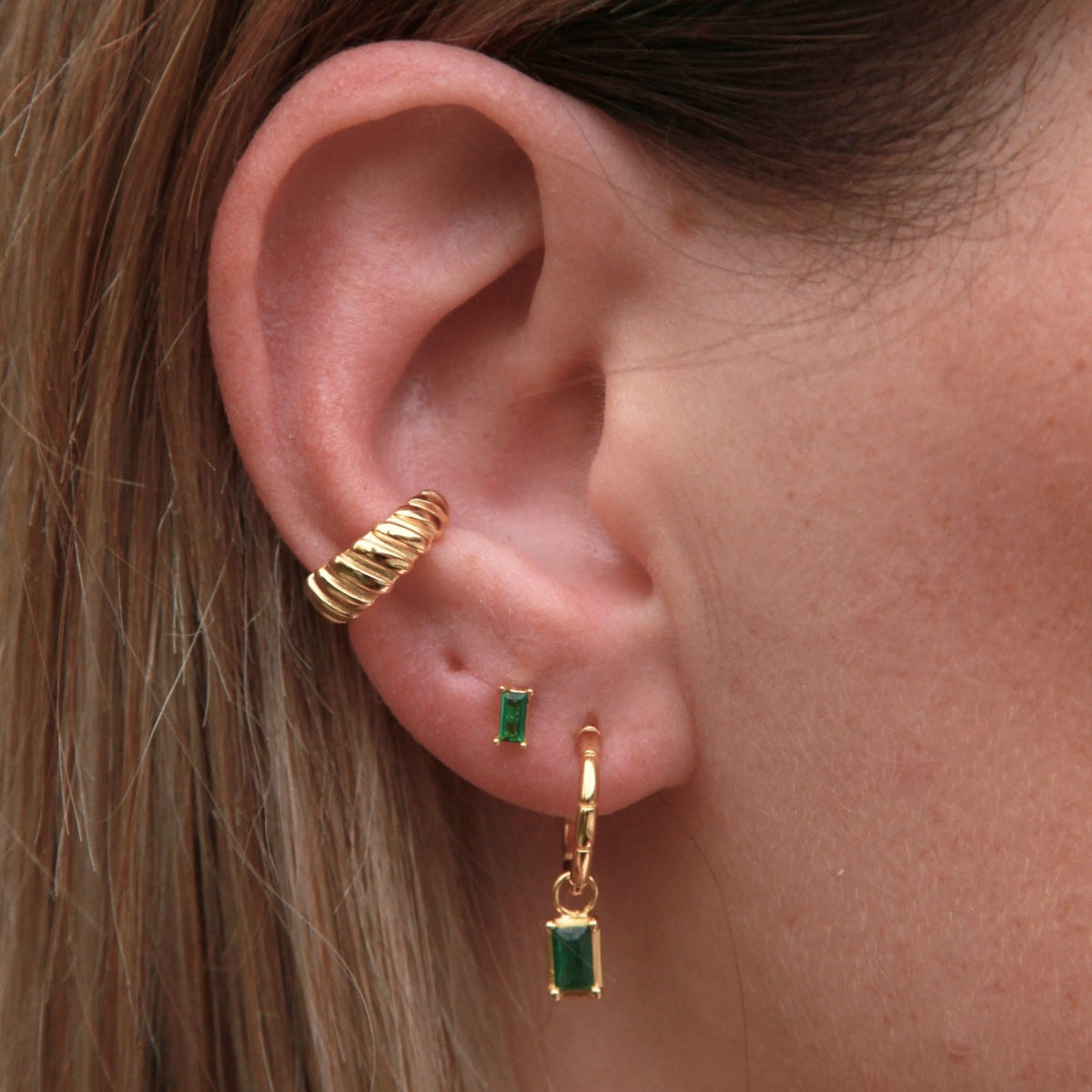 Close-up of an ear wearing gold hoop earrings with green gemstones.