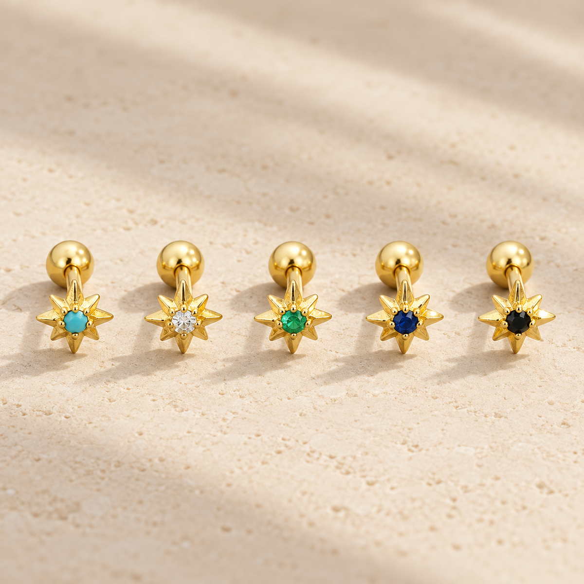 Five gold star-shaped stud earrings with round gemstone centers in light blue, clear, green, dark blue, and black, arranged on a textured beige surface with soft shadows.