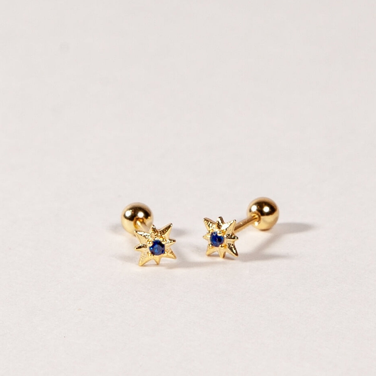 Pair of gold star shape earrings with sapphire blue CZ Stones on a light gray background