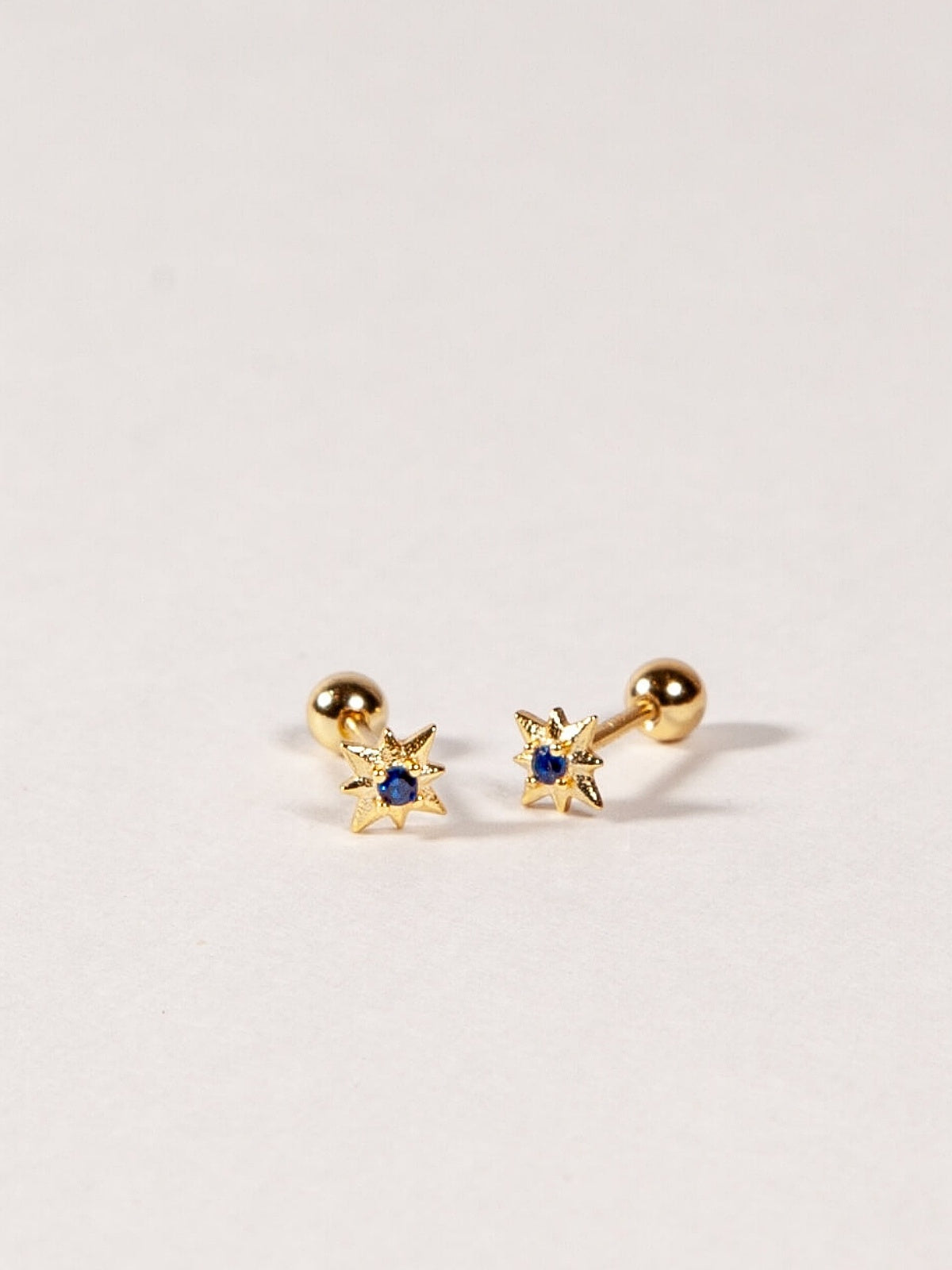 Pair of gold star shape earrings with sapphire blue CZ Stones on a light gray background