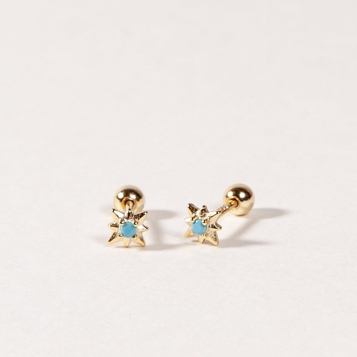 Pair of gold star shape earrings with turquoise blue CZ Stones on a light gray background