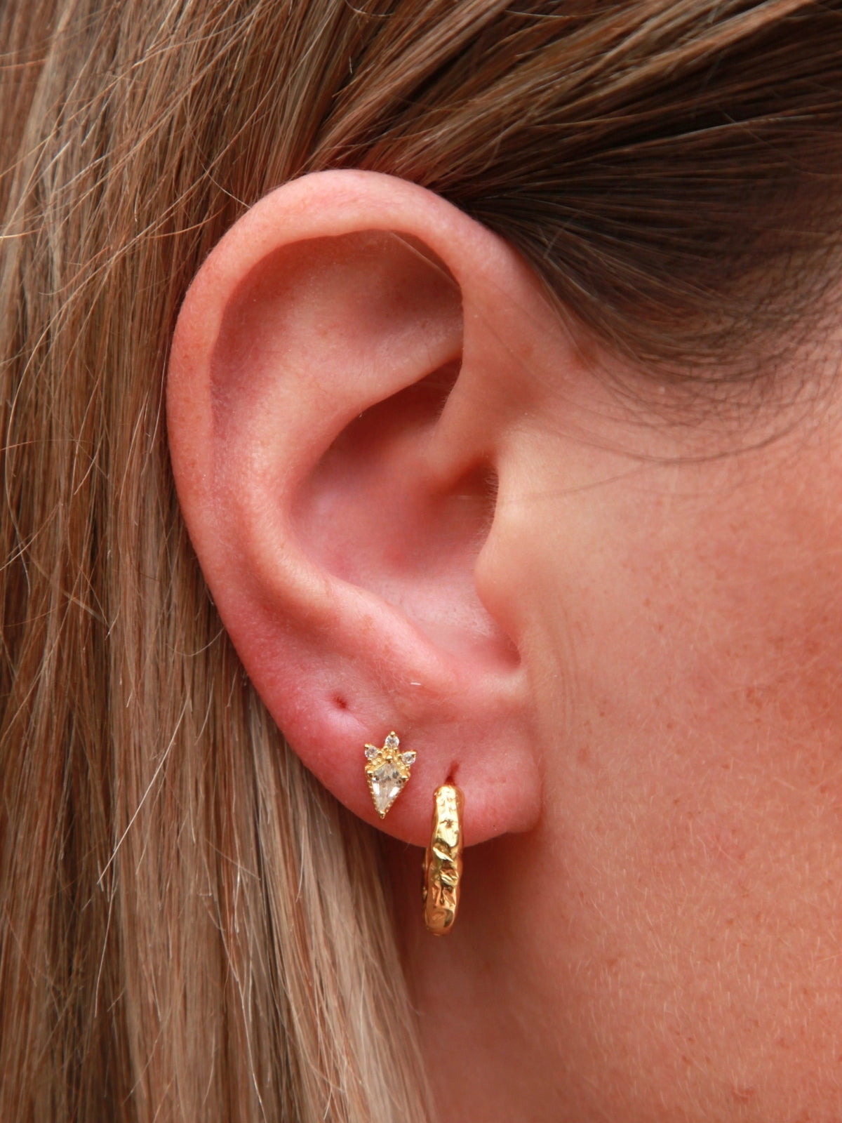 Close-up of an ear wearing gold hoop earrings and a kite shape stud.