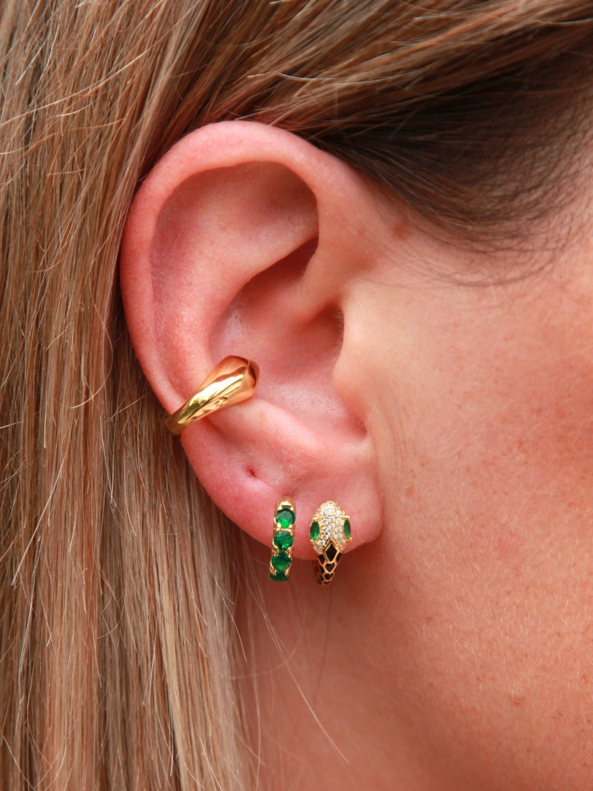 Gold snake hoop earrings with green gemstones worn by a person.